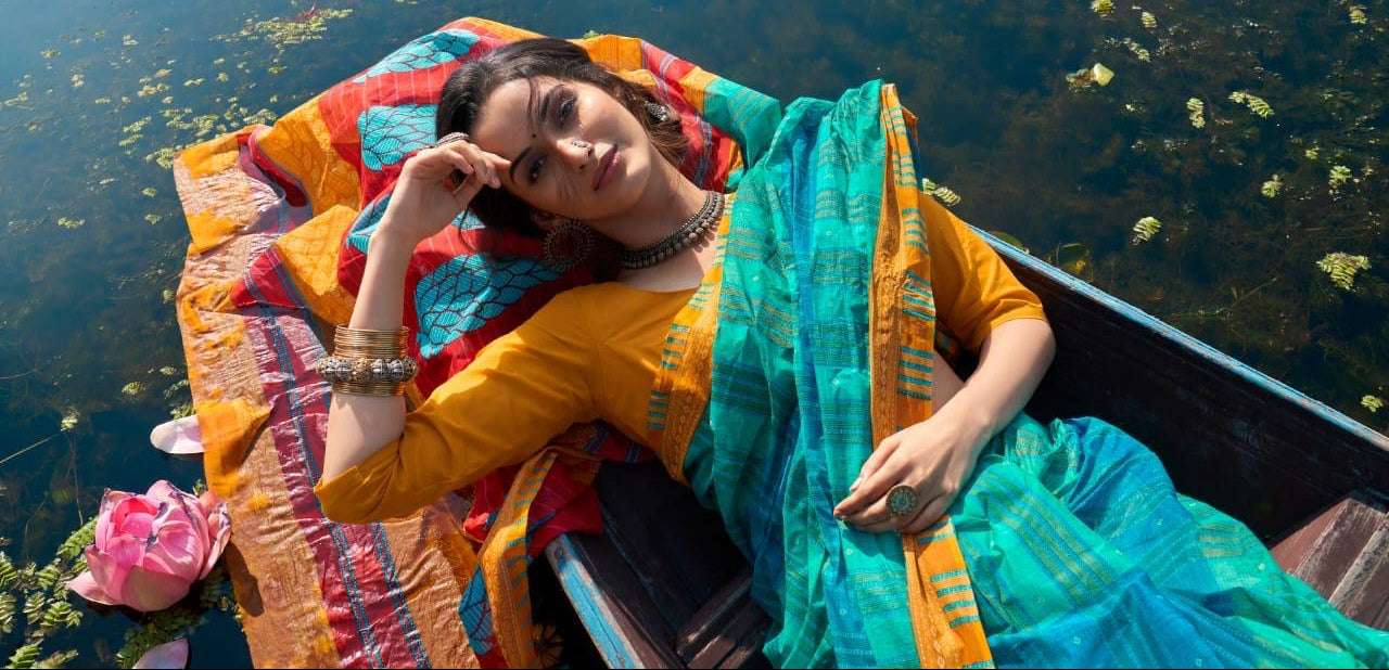 A woman reclining in a wooden boat on a lake, wearing a bright yellow kurta and a teal and orange patterned sari.
